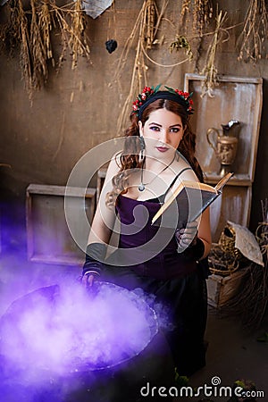 A young witch with a book in her hands cooks a potion in a large black cauldron, emitting magical purple smoke Stock Photo