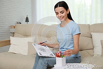 Young woman with antistress coloring page taking pencil on sofa in room Stock Photo