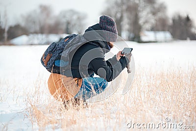 A young woman with a backpack on her back, squats down and takes pictures of dry grass on her phone. Side view Stock Photo