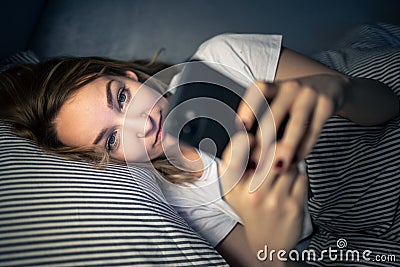 Young woman in bed holding a phone, tired and exhausted, blue light straining her eyes Stock Photo