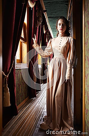 Young woman in beige vintage dress of early 20th century standing near window in corridor of retro railway train Stock Photo