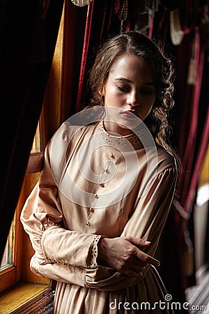 Young woman in beige vintage dress of early 20th century standing near window in corridor of retro railway train Stock Photo