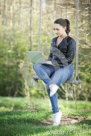 Young woman in a black jacked with coffee cup and laptop working outside in a park. Remote work. Digital work. Stock Photo