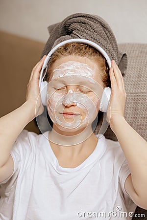 Young woman with casual clothes, towel on head and beauty mask on face sitting on couch at home, resting and listening Stock Photo