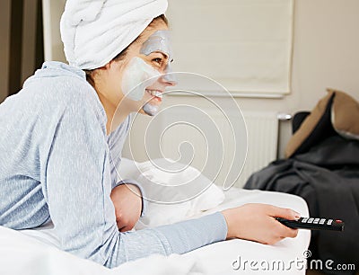 Young woman with clay mask on face watching tv at home. Stock Photo