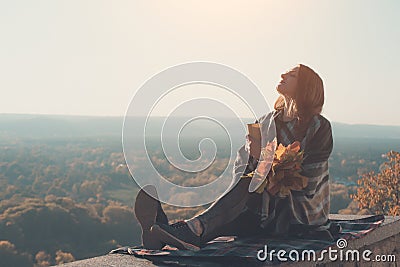 Young woman with closed eyes sits on a hill. Paper cup in hands and a bouquet of yellow leaves. Enjoying a sunny day Stock Photo