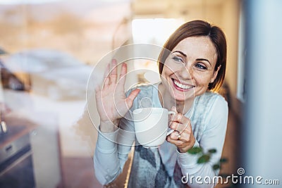 A young woman with cup of coffee looking out of a window, waving goodbye. Stock Photo