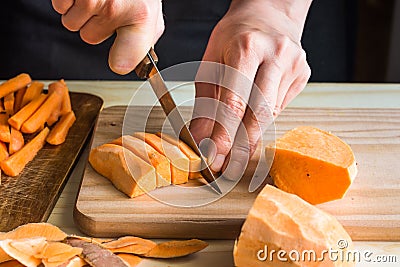 Young woman cutting with knife sweet potato into wedges, peels on wood table, sliced carrots Stock Photo