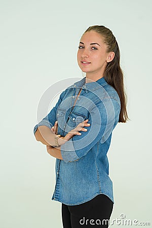 Young Woman in Denim Shirt and Jeans, Leaning Against Gray Wall Background with Arms Crossed and Looking at the Camera Stock Photo