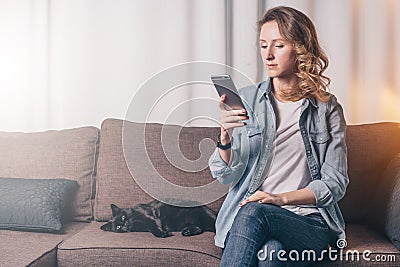 Young woman in denim shirt sitting at home on couch and using smartphone,nearby lies cat. Girl uses digital gadget. Stock Photo