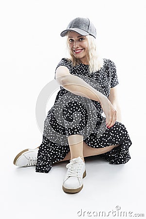 A young woman in a dress is sitting. Smiling stylish blonde in a black dress and a cap. White background. Vertical Stock Photo