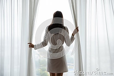 Young woman dressed in bathrobe opening curtains on picture window Stock Photo