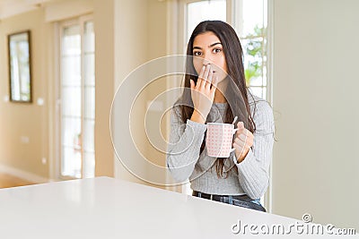 Young woman drinking a cup of coffee cover mouth with hand shocked with shame for mistake, expression of fear, scared in silence, Stock Photo