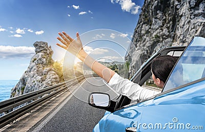 A young woman is driving by car to the sea and waving her hand from a blue convertible car. Vacation on the sea coast. Stock Photo