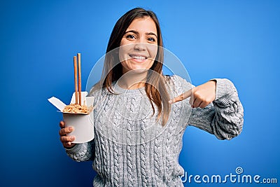 Young woman eating asian noodles from take away box using chopstick over blue background with surprise face pointing finger to Stock Photo