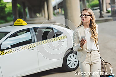 young woman in eyeglasses holding smartphone and looking away while standing near taxi cab Stock Photo