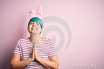 Young woman with fashion blue hair wearing easter rabbit ears over pink background begging and praying with hands together with Stock Photo