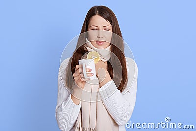 Young woman fell ill, she got sick and sore throat, keeping eyes closed, touching her neck, drinking hot tea with lemon, wearing Stock Photo