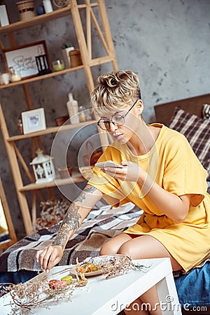 Food Blogger. Young woman in glasses sitting at stylish apartment taking photo of desserts on plate on smartphone Stock Photo