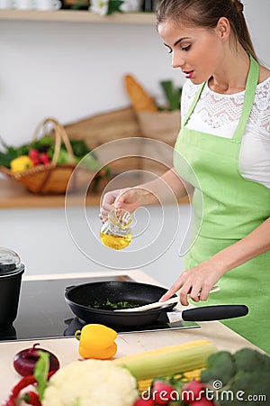 Young woman in the green apron cooking in the kitchen. Housewife cooks the meal in a frying pan . Stock Photo