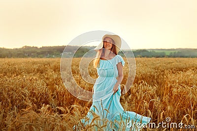 A young woman in a hat and a blue dress is spinning in a wheat field. The lady enjoys the sunset and nature Stock Photo