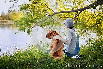 Young woman in a hat with dog Shar Pei sitting in the field and looking to the river in golden sunset light Stock Photo