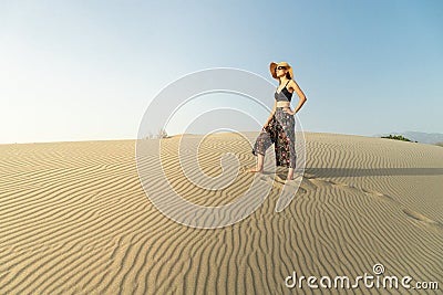 Young woman with hat walking in the desert dunes with footsteps in the sand during sunset Stock Photo