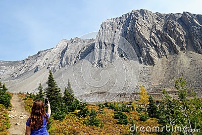 A young woman hiking and taking a selfie photo by a beautiful trail with a huge mountain in the background during a day in autumn Stock Photo