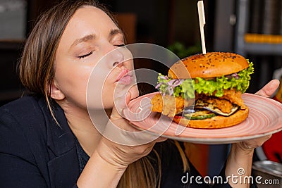 Young woman hold in hands plate with juicy appetizing chicken burger and enjoy flavour with closed eyes. Serving of big Stock Photo