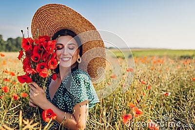 Young woman holding bouquet of poppies flowers walking in summer field. Stylish happy girl wearing straw hat Stock Photo
