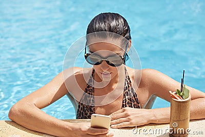 Young woman holding phone in hand and checking social networks while drinking cocktail at edge of swimming pool. Female in Stock Photo