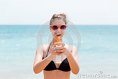 Young woman is holding a spray of sun cream for body at the camera at the beach Stock Photo