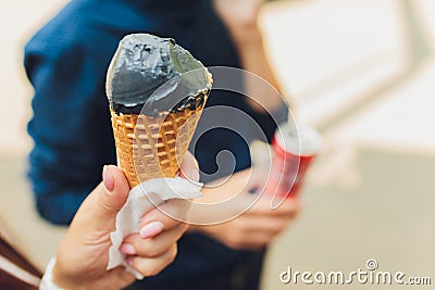 Young woman holds in hands dark black cone ice-cream. Close-up portrait female with red lips. Black waffle horn. Trendy Stock Photo