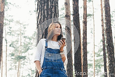Young woman in jeans overalls with smartphone standing in woodland Stock Photo