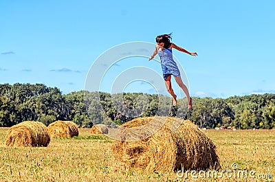 Young woman jumps on haystack in a field. Her hands apart. Blue sky and green trees at background. Large bundles of hay Stock Photo