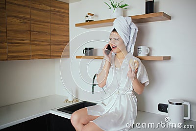 Young woman in kitchen during quarantine. Girl after shower sit and talk on phone. Hold tasty donut in hand Stock Photo