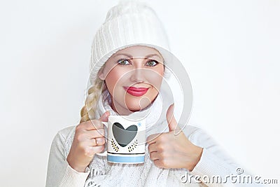 A young woman in a knitted hat and scarf holding a mug of tea and showing her thumb up. Stock Photo