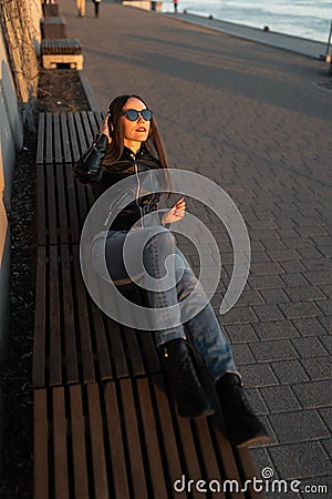 Young woman listens to music in closed headphones through her phone wearing a leather jacket and jeans at a sunset near Stock Photo