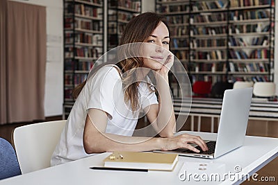 Young woman long hair analyst working at co-working office on laptop, look to camera, calm face, bookshelf behind, library Stock Photo