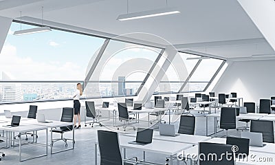 A young woman is looking out the window in the modern bright open space loft office. White tables equipped by modern laptops and b Stock Photo