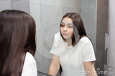 Young woman looks in the mirror. Girl with long dark hair in the bathroom looks at herself in the mirror. Stock Photo