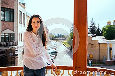 A young woman looks out from the balcony.The bright sun illuminates the girl. Beautiful brunette enjoys the sun Stock Photo