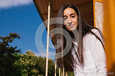 A young woman looks out from the balcony.The bright sun shines on the girl. Beautiful brunette girl enjoys the sun Stock Photo