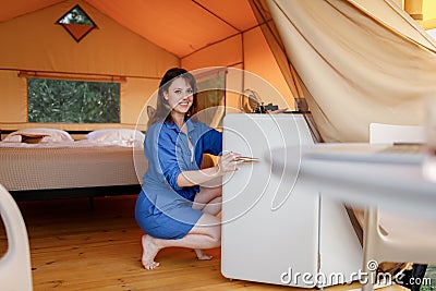 Young woman looks into the refrigerator during vacation in modern glamping tent. Luxury camping tent for outdoor recreation Stock Photo