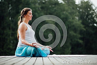 Young woman in the lotus position is practicing yoga in the forest next to the river. sitting on mats the wooden pier. Stock Photo