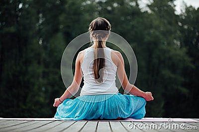 Young woman in the lotus position is practicing yoga in the forest next to the river. sitting on mats the wooden pier. Stock Photo