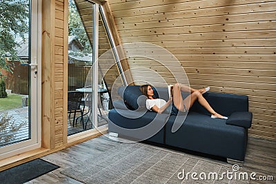 Young woman lying on couch inside cabin in woods relaxing with a book Stock Photo