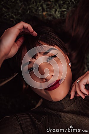 A young woman is lying on the grass, showing off her beautiful face with a serene expression. The lush greenery in the Stock Photo