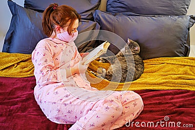 A young woman in a makeshift medical mask lies with the pet during the epidemic of the coronavirus. A girl in pajamas on the bed Stock Photo