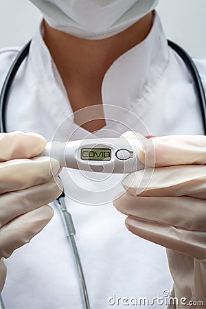A young woman medic holds an electronic thermometer in her hands which shows the word COVID on the display. The concept of Stock Photo